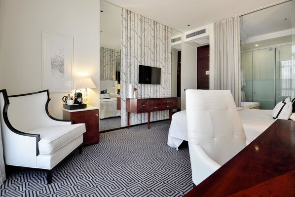 A modern hotel room with a large bed, two white armchairs, a wooden desk, a TV mounted on a wall, and a patterned floor.