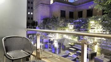 A balcony with a checkered floor, wicker furniture, and a view of a building with illuminated windows.