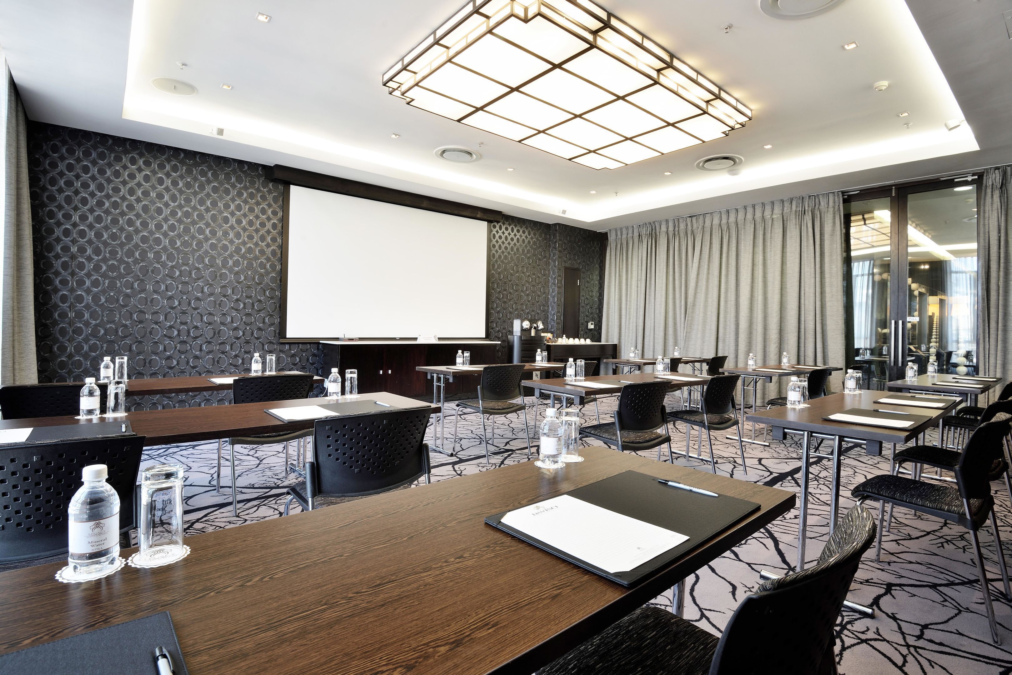 Uma sala de conferências com mesas, cadeiras, uma tela de projetor e um grande janelão com cortinas.
