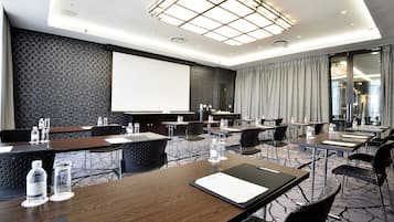Uma sala de conferências com mesas, cadeiras, uma tela de projetor e um grande janelão com cortinas.
