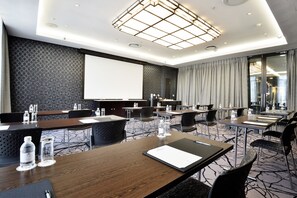 Un salón de conferencias con mesas, sillas, una pantalla de proyector y un amplio ventanal con cortinas.