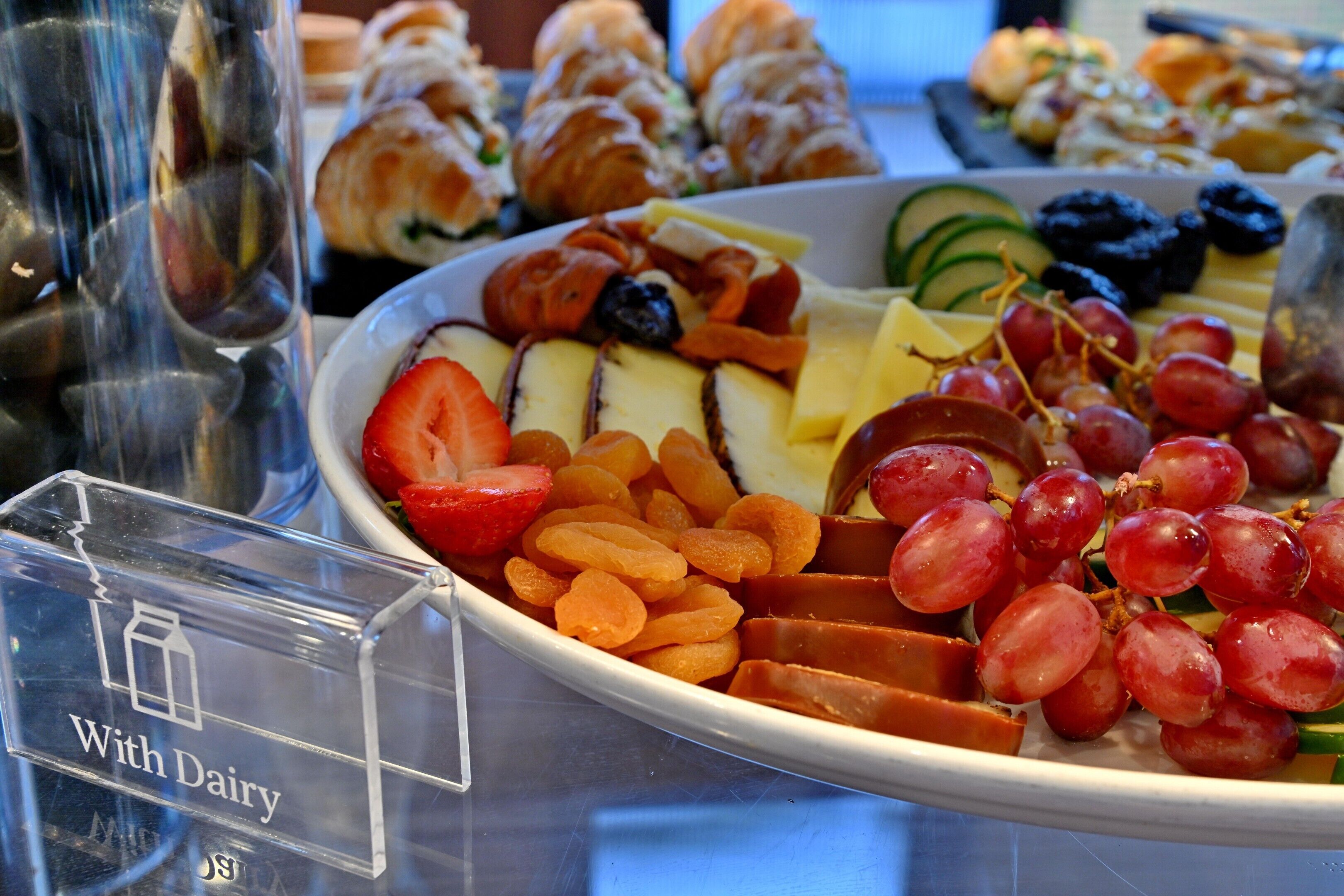 A platter of assorted fruits and cheeses, including grapes, strawberries, and cheese slices, with a sign indicating 'With Dairy.'
