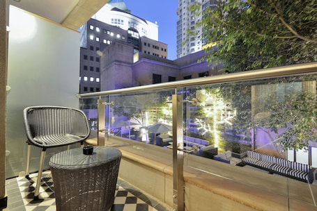 A balcony with a wicker chair and table, a glass railing, and a view of a cityscape and greenery.