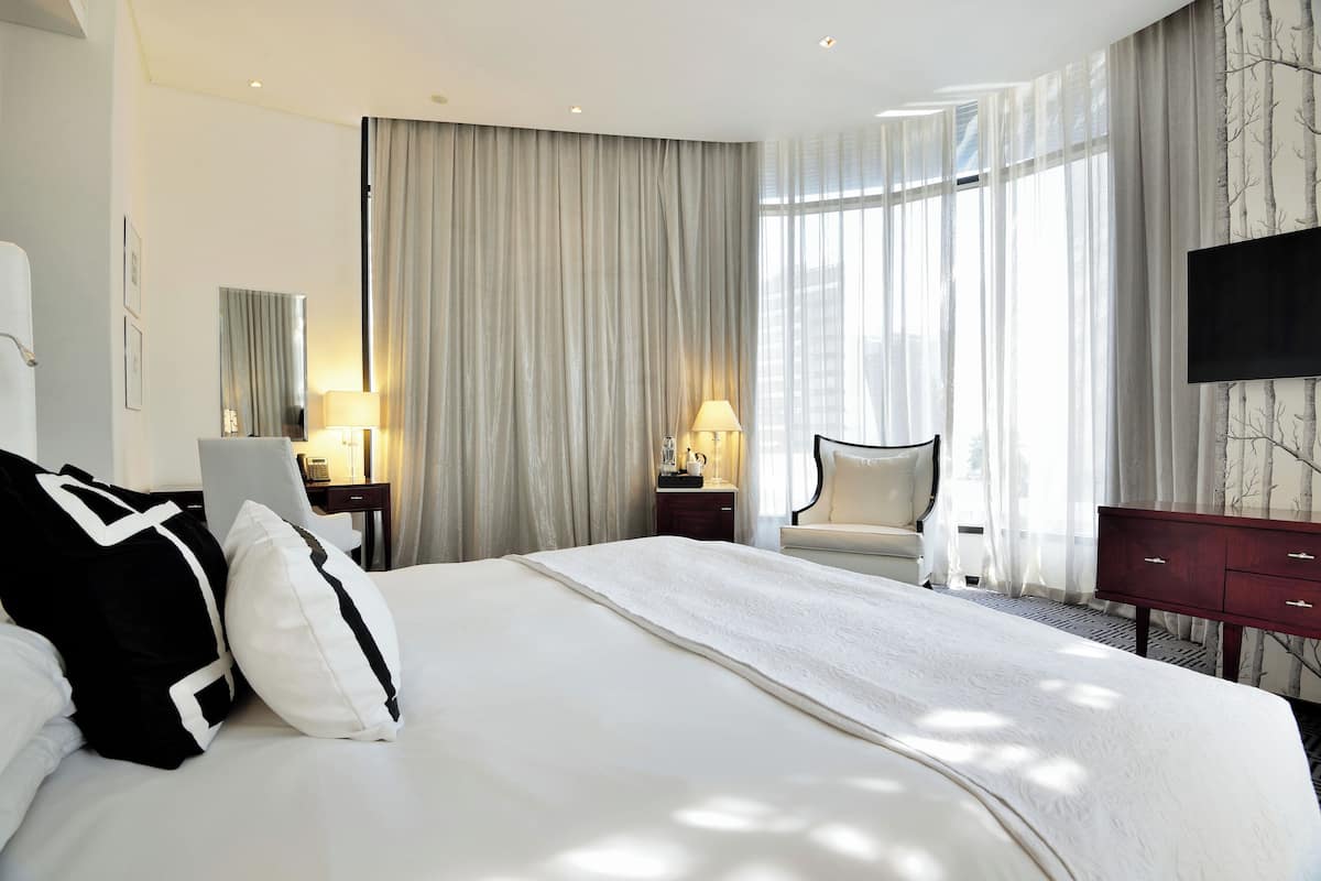 A hotel room with a large bed, a desk with a lamp, a chair, and a TV on the wall.