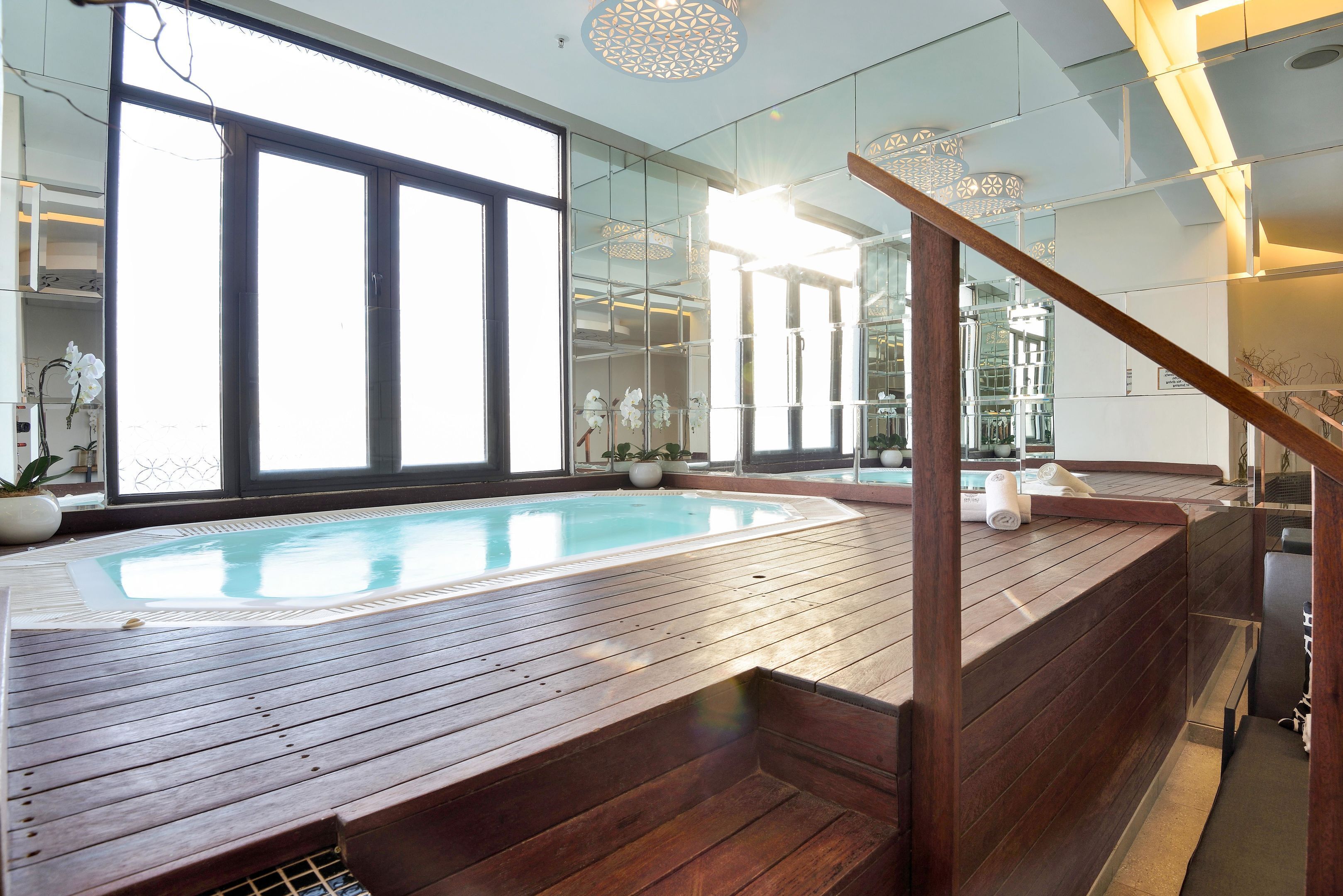 Una piscina interior moderna con cubierta de madera, ventanas amplias y una pared de vidrio que refleja la piscina.