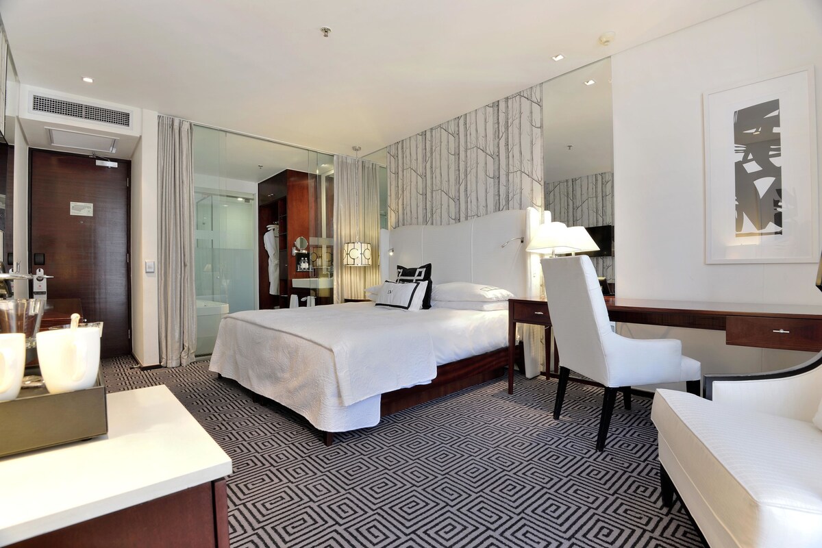 A modern hotel room with a large bed, a desk, a chair, a bedside table, a mirror, and a bathroom with a shower and toilet.