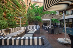 Un área de estar al aire libre con sofás a rayas y sombrillas, rodeada de vegetación y una piscina.