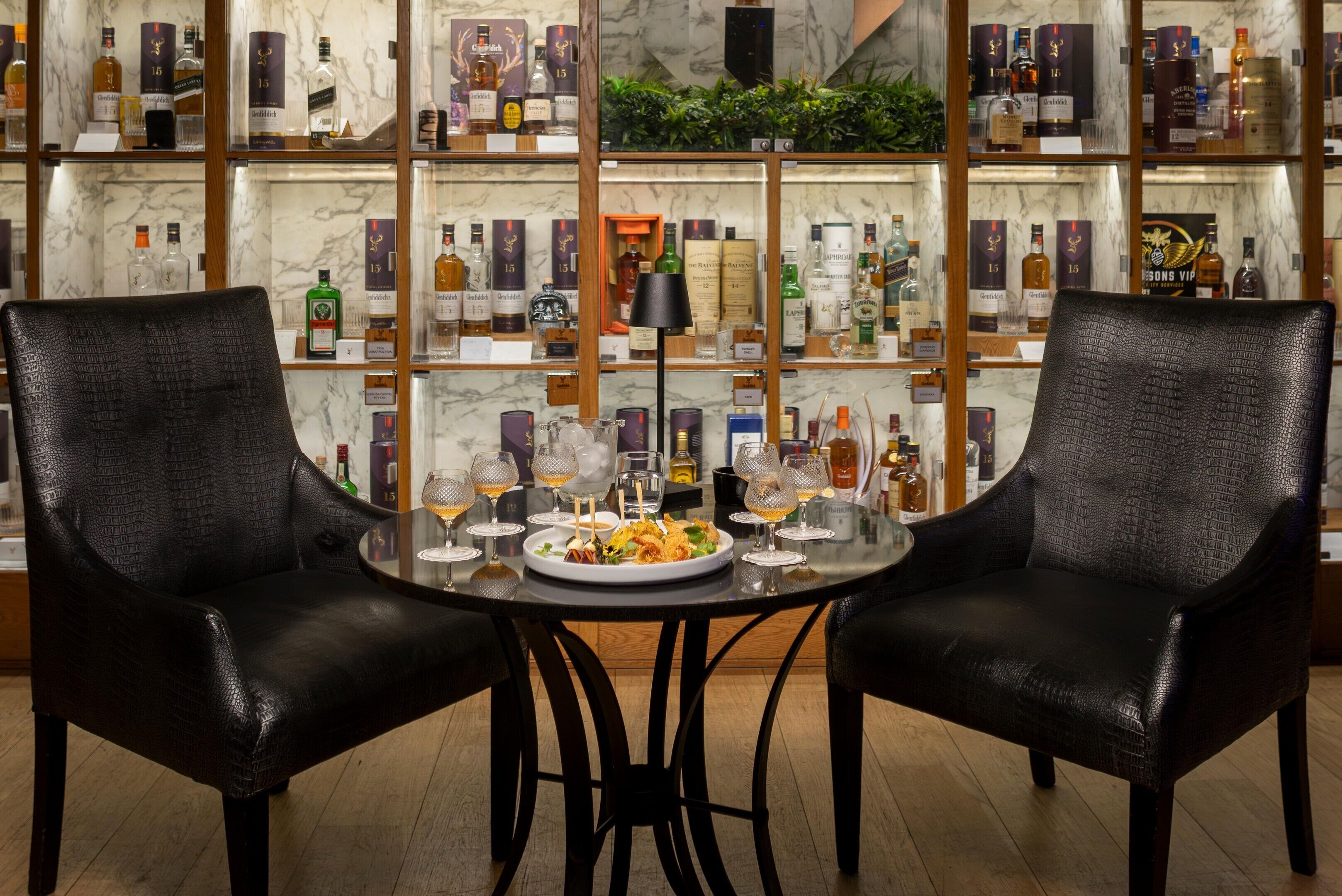 Una zona de bar con dos sillones de cuero negro, una mesa redonda con un plato de comida y vasos, y estantes llenos de botellas de licores.