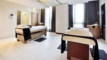Una habitación de hotel moderna con dos camas, un espejo grande, un banco de madera y un pequeño escritorio con una lámpara.