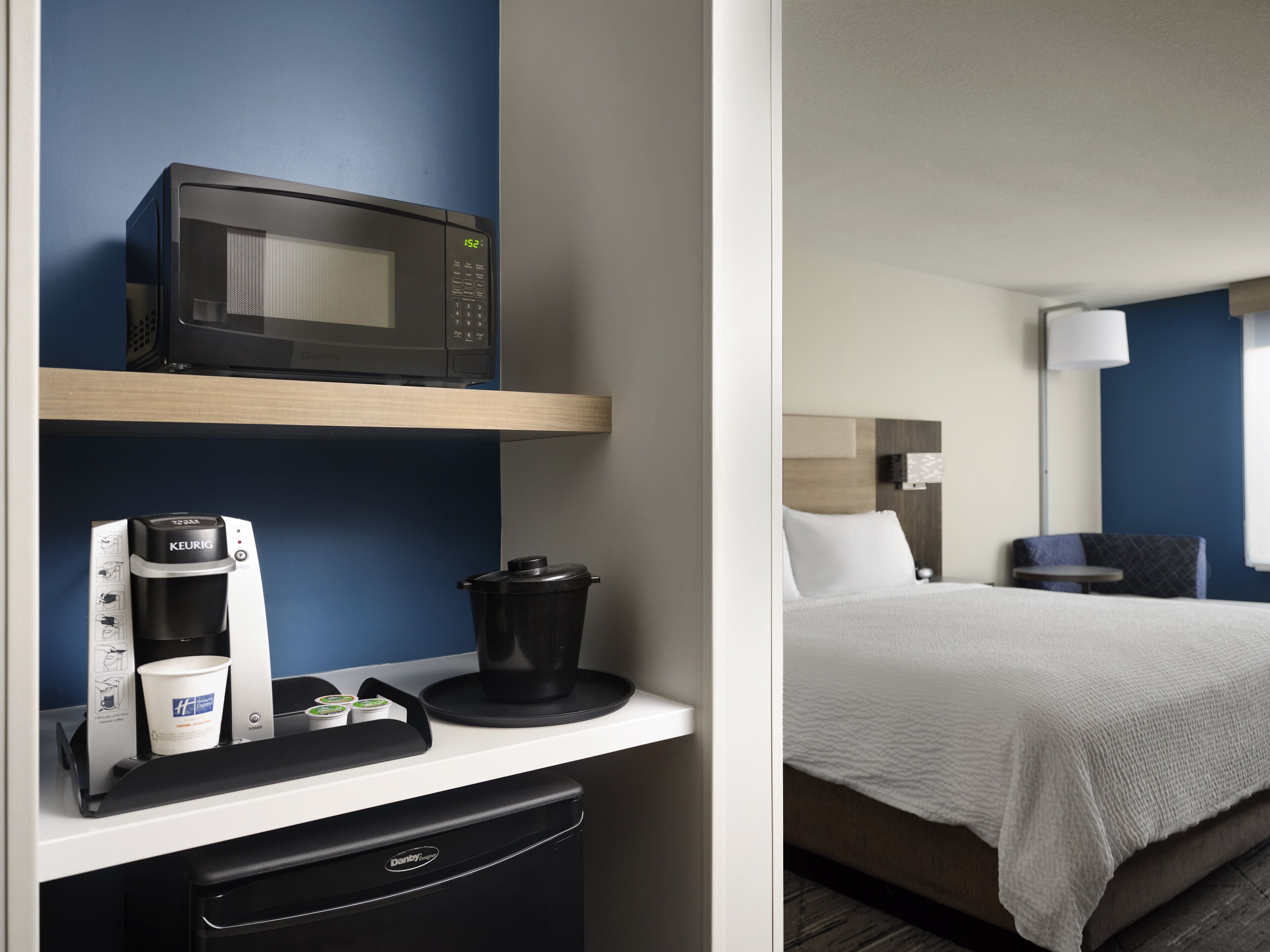 A hotel room with a microwave on a shelf, a coffee maker, a pot, and a bed with a white comforter.