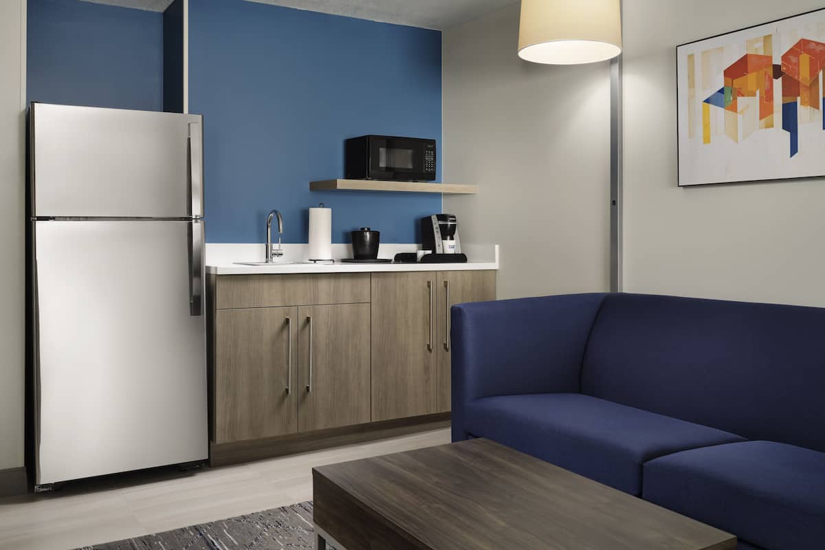 Una cucina moderna con frigorifero, forno a microonde e macchina del caffè su un piano di lavoro bianco. Nell'area soggiorno, un divano blu e un tavolino in legno.