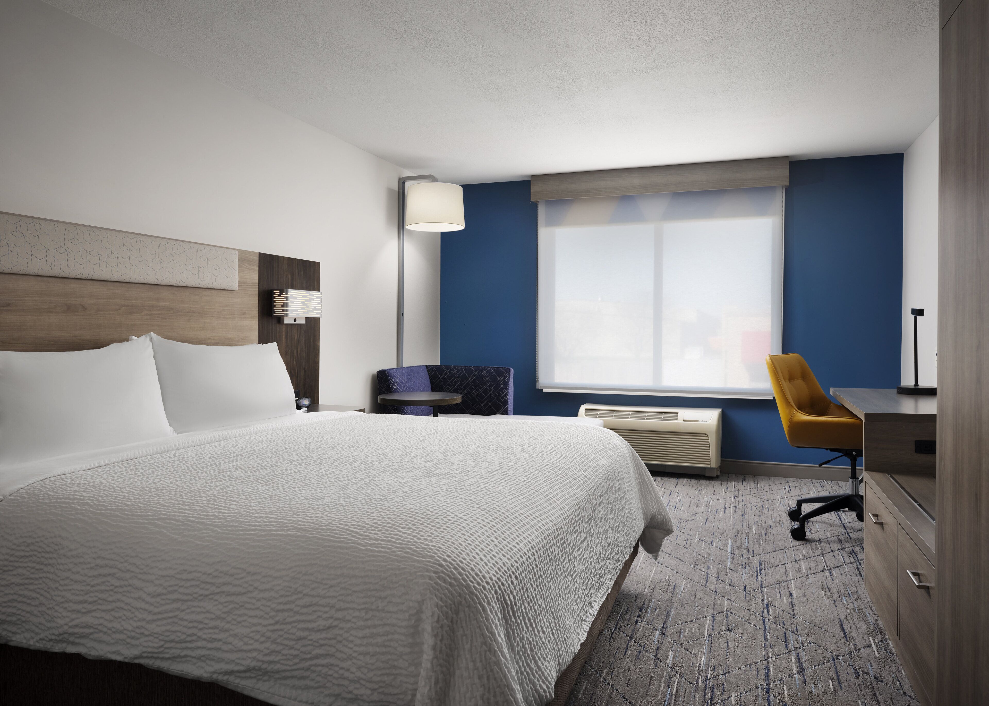 A hotel room with a bed, a desk, a chair, a window, and an air conditioning unit.