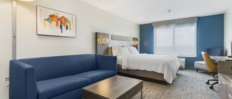A modern hotel room with a blue sofa, a wooden coffee table, a bed with white linens, and a desk with a chair.