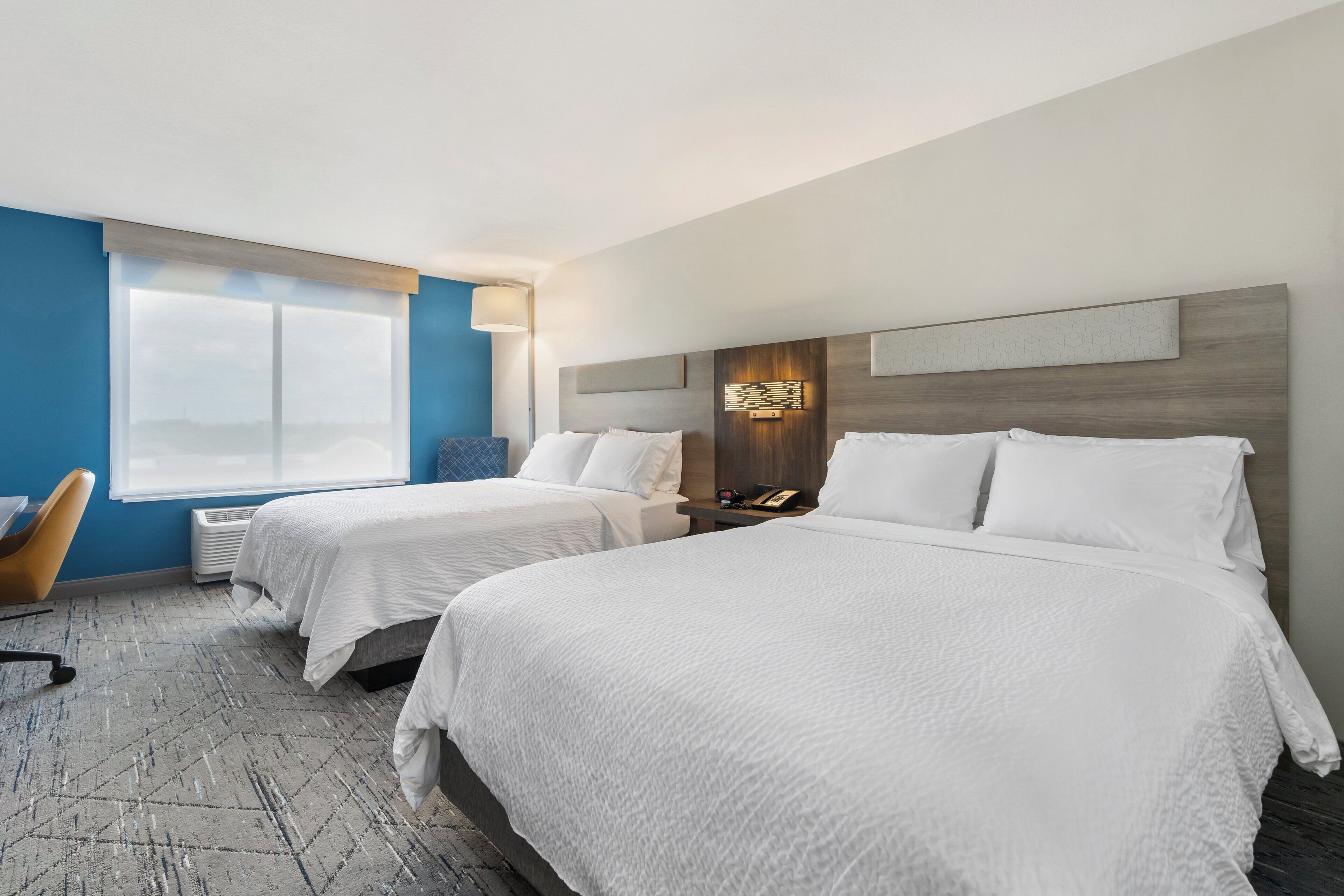 Quarto de hotel com duas camas, uma escrivaninha, uma cadeira e uma janela grande.