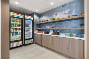 A clean, modern kitchenette with two refrigerators stocked with beverages, shelves with snacks, and a countertop with various kitchen items.