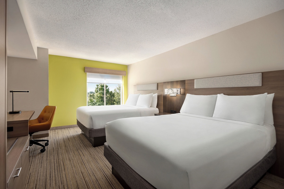 A hotel room with two beds, a desk, a chair, and a window with a view of trees.