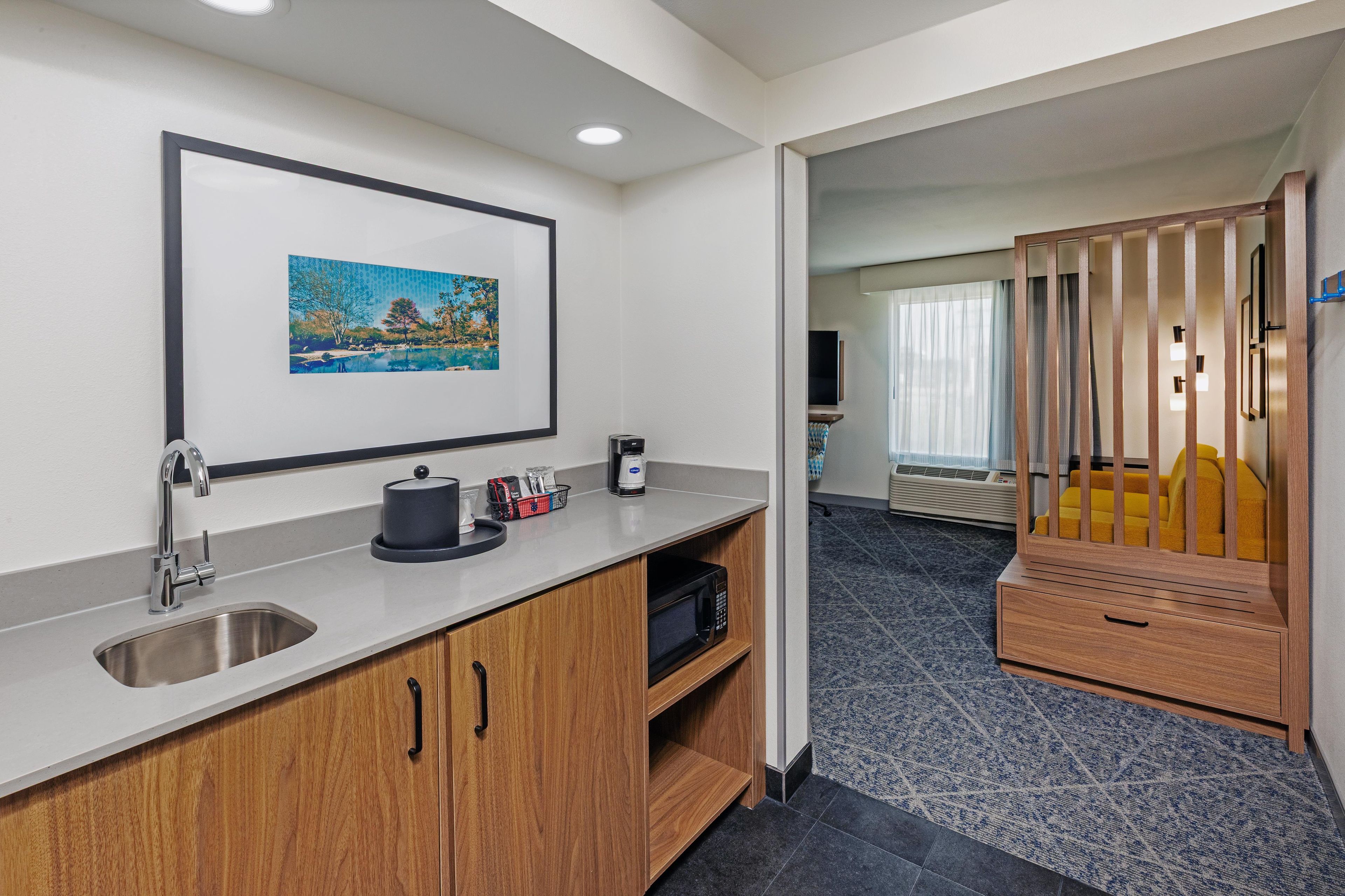 A hotel room with a kitchenette, a framed picture on the wall, a wooden cabinet, and a view into the living area.