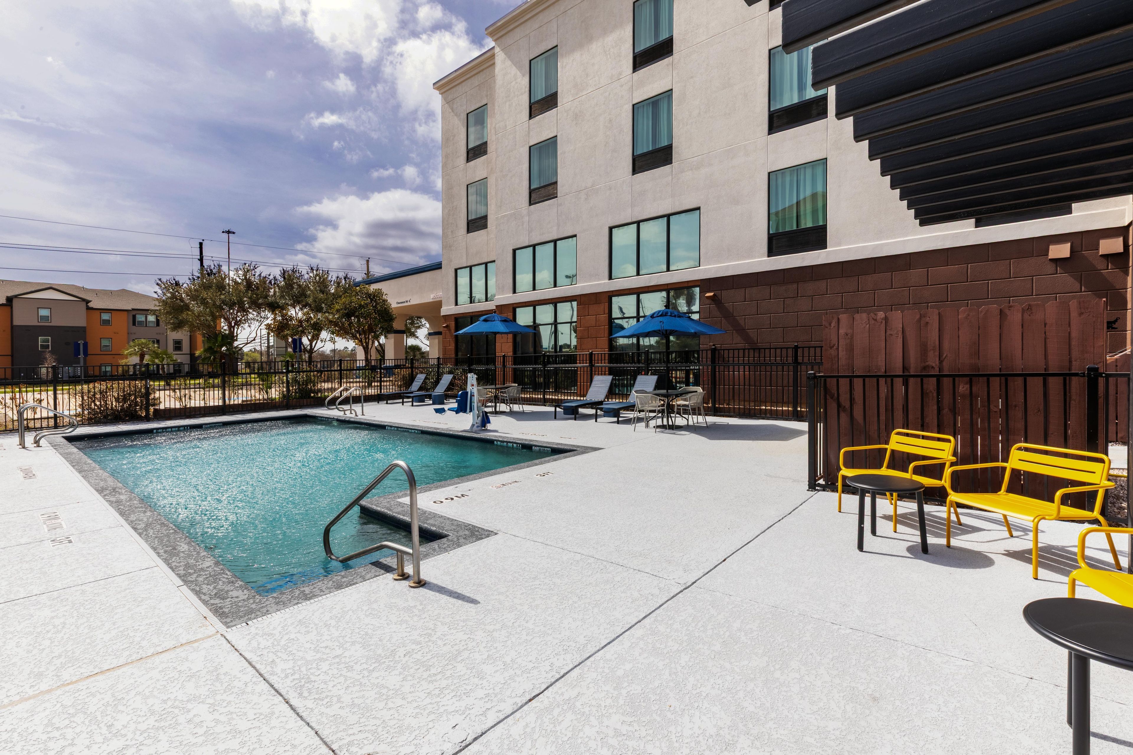 A swimming pool with a metal ladder, surrounded by lounge chairs and umbrellas, adjacent to a building with multiple windows and a brick facade.