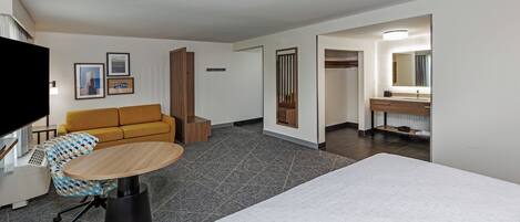 A hotel room with a bed, a sofa, a dining table, a TV, a bathroom with a mirror and sink, and framed pictures on the wall.