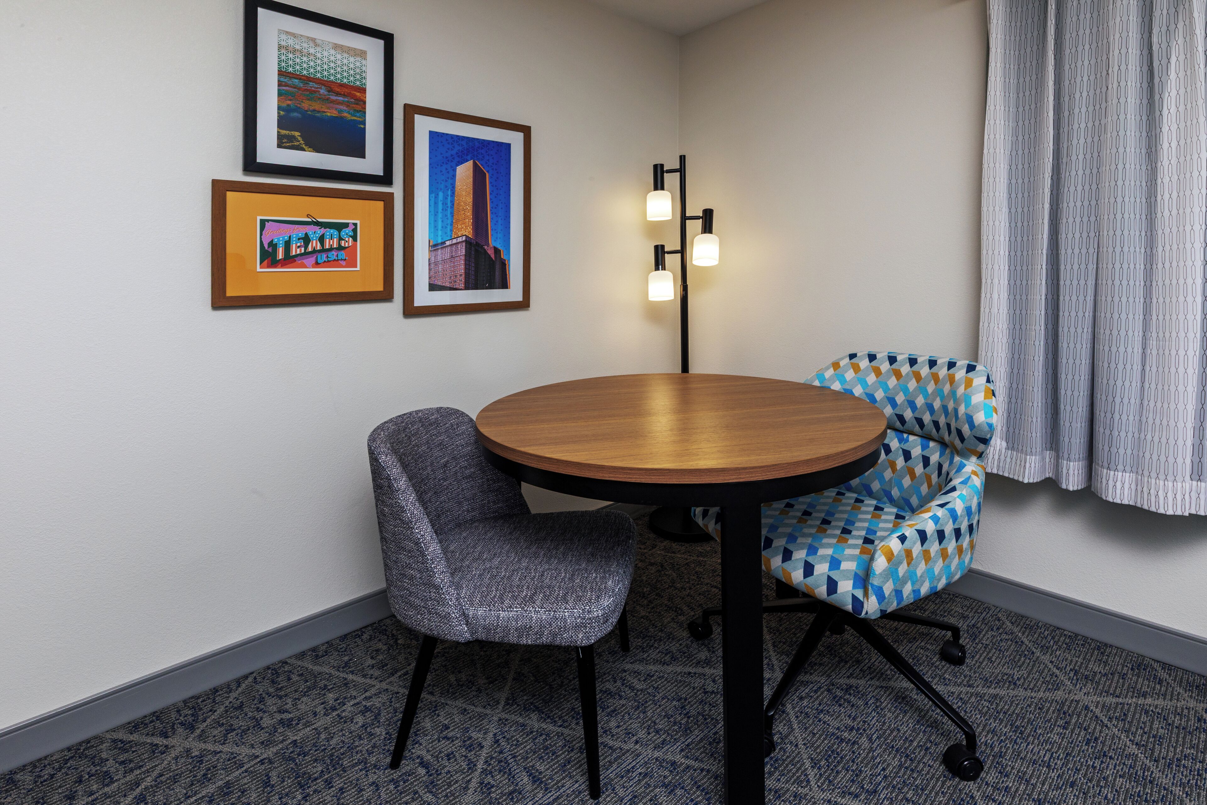 A conference room with a round table, two chairs, framed pictures on the wall, and a floor lamp.