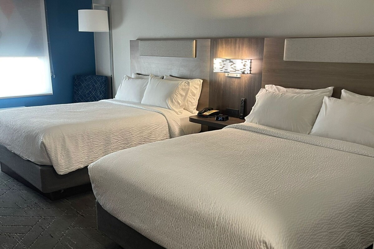 Hotelkamer met twee bedden, een nachtkastje met telefoon en wekker, een wandlamp en een raam met jaloezieën.