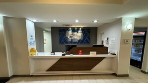 A hotel reception area with a counter, a digital screen, a refrigerator with beverages, and a blue wall with geometric patterns.