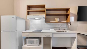 Una cocina compacta con electrodomésticos blancos, un microondas y un lavamanos. Hay estantes de madera sobre la encimera y un televisor de pantalla plana montado en la pared.