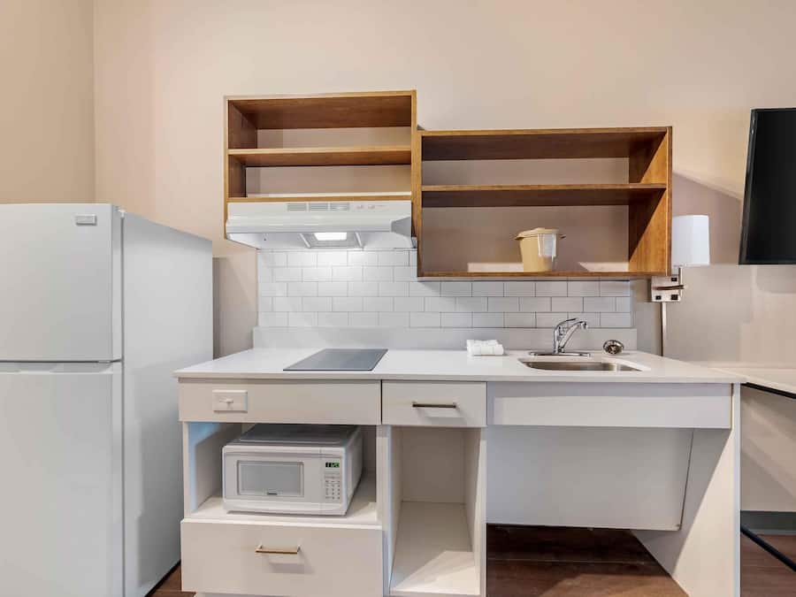 A compact kitchen with white appliances, a microwave, and a sink. There are wooden shelves above the counter and a flat-screen TV mounted on the wall.