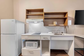A compact kitchen with white appliances, a microwave, and a sink. There are wooden shelves above the counter and a flat-screen TV mounted on the wall.