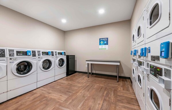 A laundry facility with multiple washing machines and dryers, a table, and a sign promoting an app. - Extended Stay America Premier Suites - San Antonio - North (San Antonio)