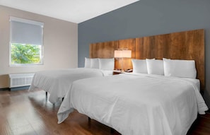 A hotel room with two beds, a wooden headboard, a window with blinds, and a bedside table with a lamp.