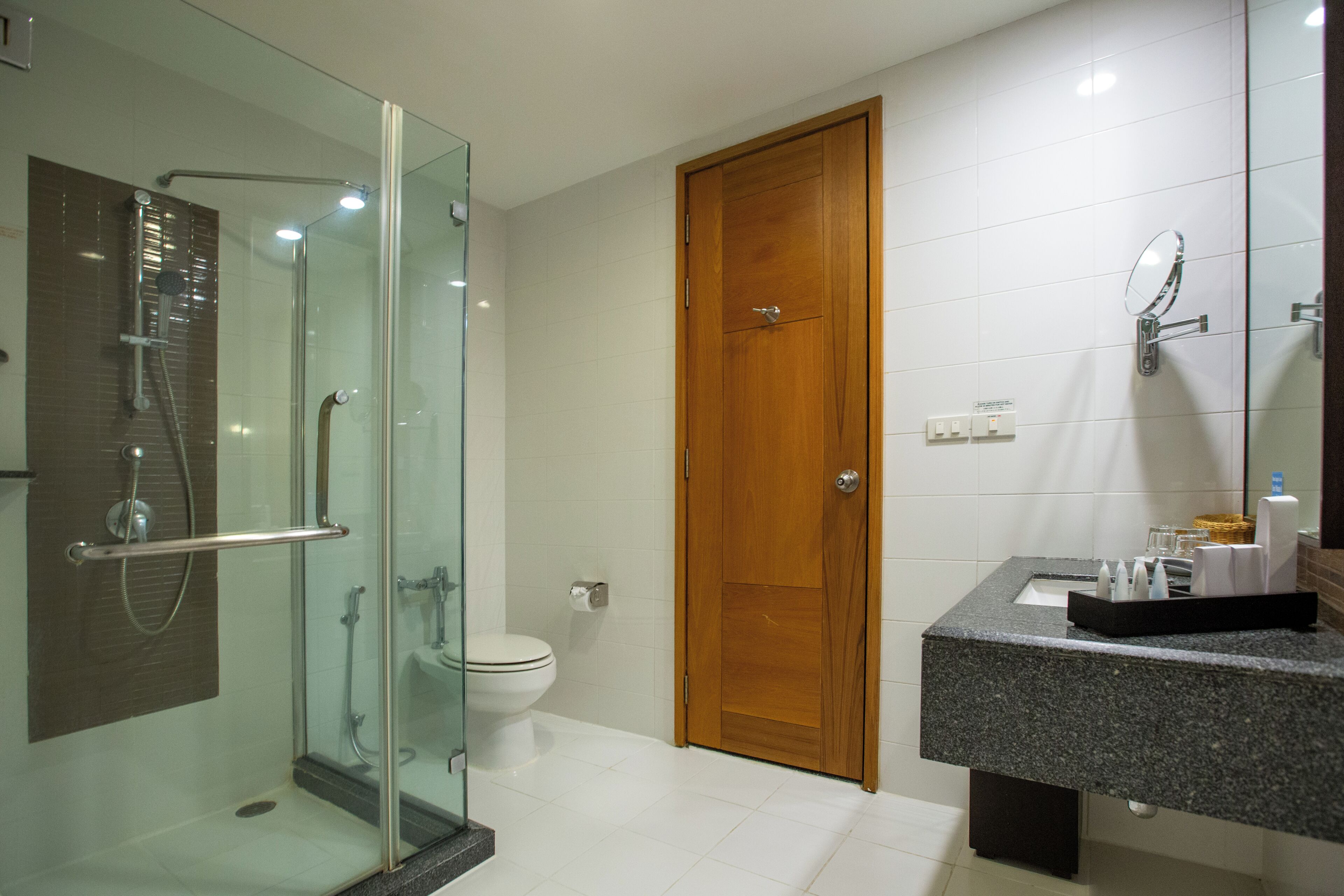 Een moderne badkamer met een douchecabine van glas, een houten deur en een granieten aanrechtblad met een spiegel en toiletartikelen.