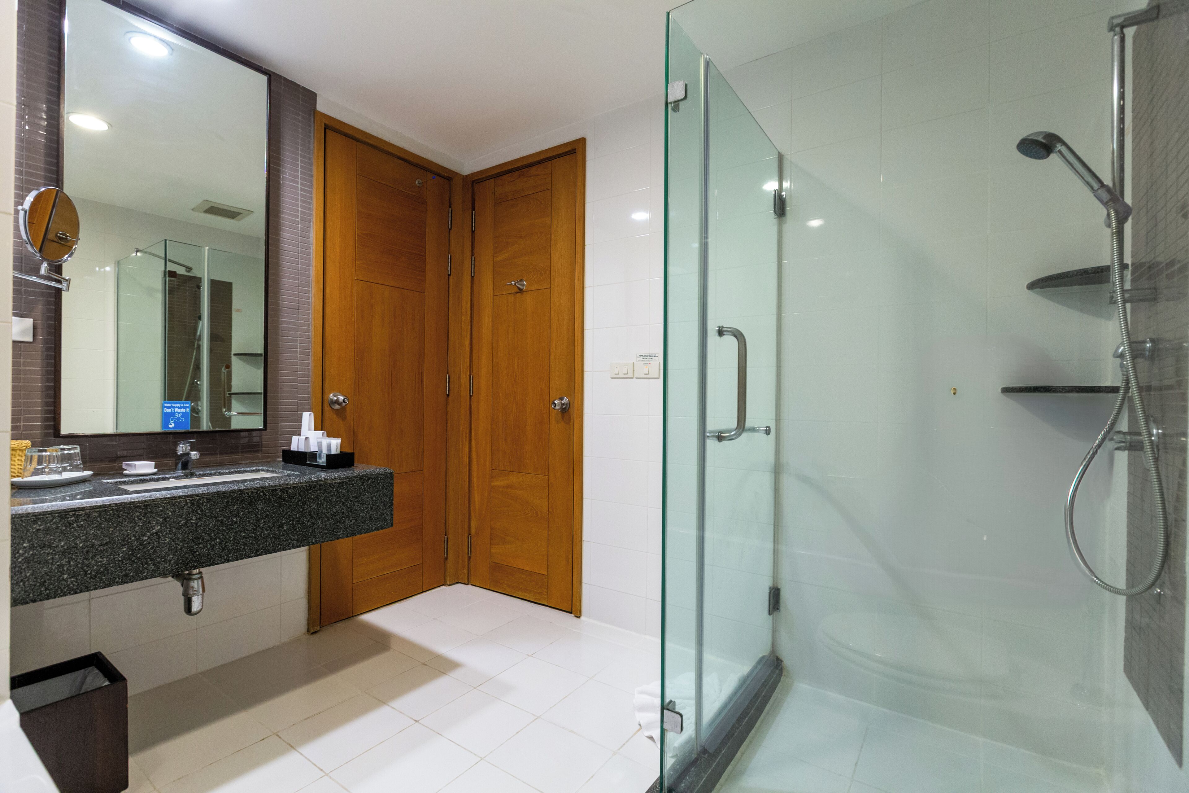 A bathroom with a glass-enclosed shower, a wooden door, a sink with a mirror, and a trash bin.