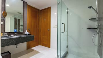 A bathroom with a glass-enclosed shower, a wooden door, a sink with a mirror, and a trash bin.