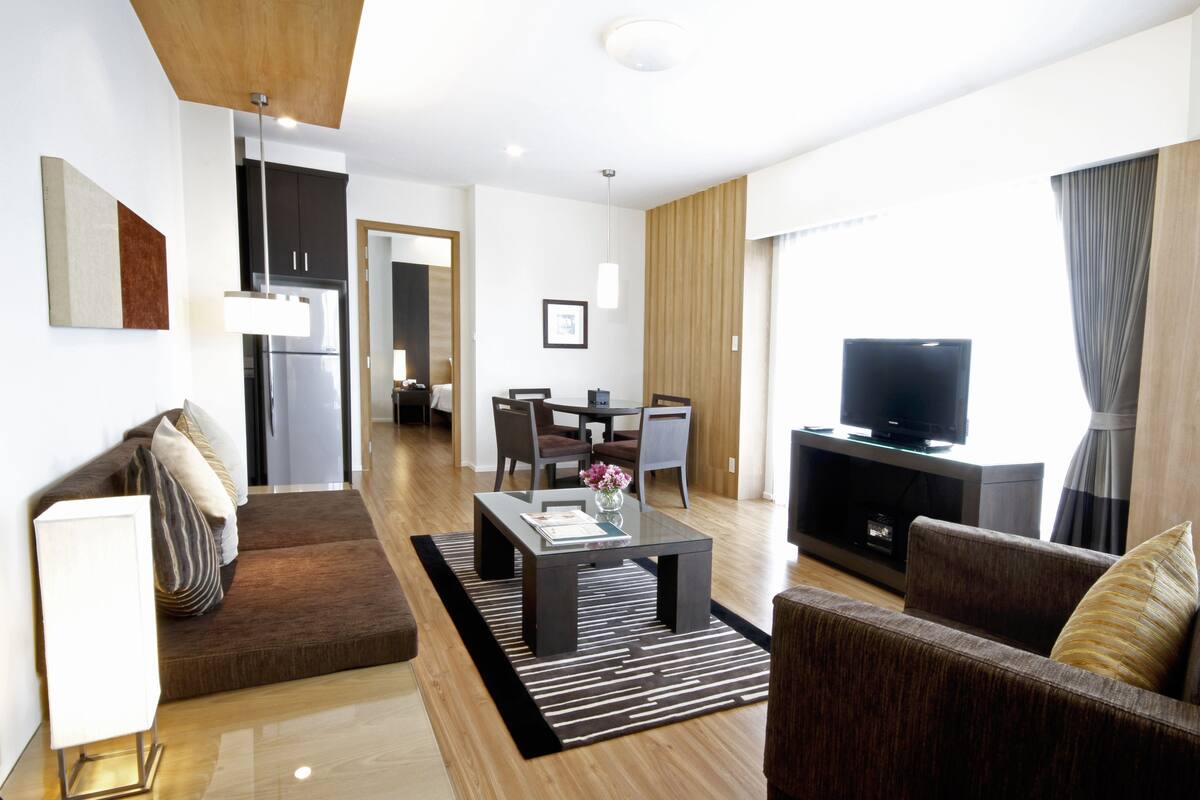 Ein modernes Wohnzimmer mit Sofa, Couchtisch und Fernseher. Es gibt einen Essbereich mit Stühlen und Tisch. Der Raum hat Holzböden und weiße Wände.