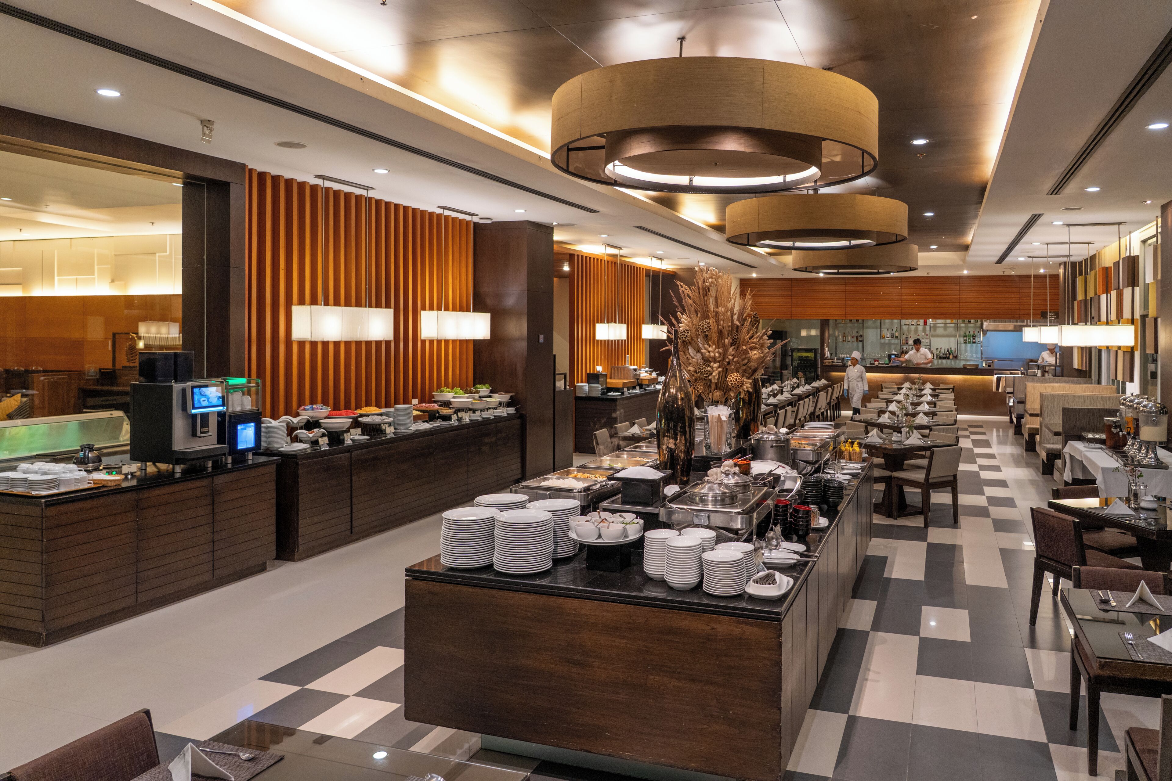 A hotel dining area with buffet service, multiple dining tables, and a central serving station.