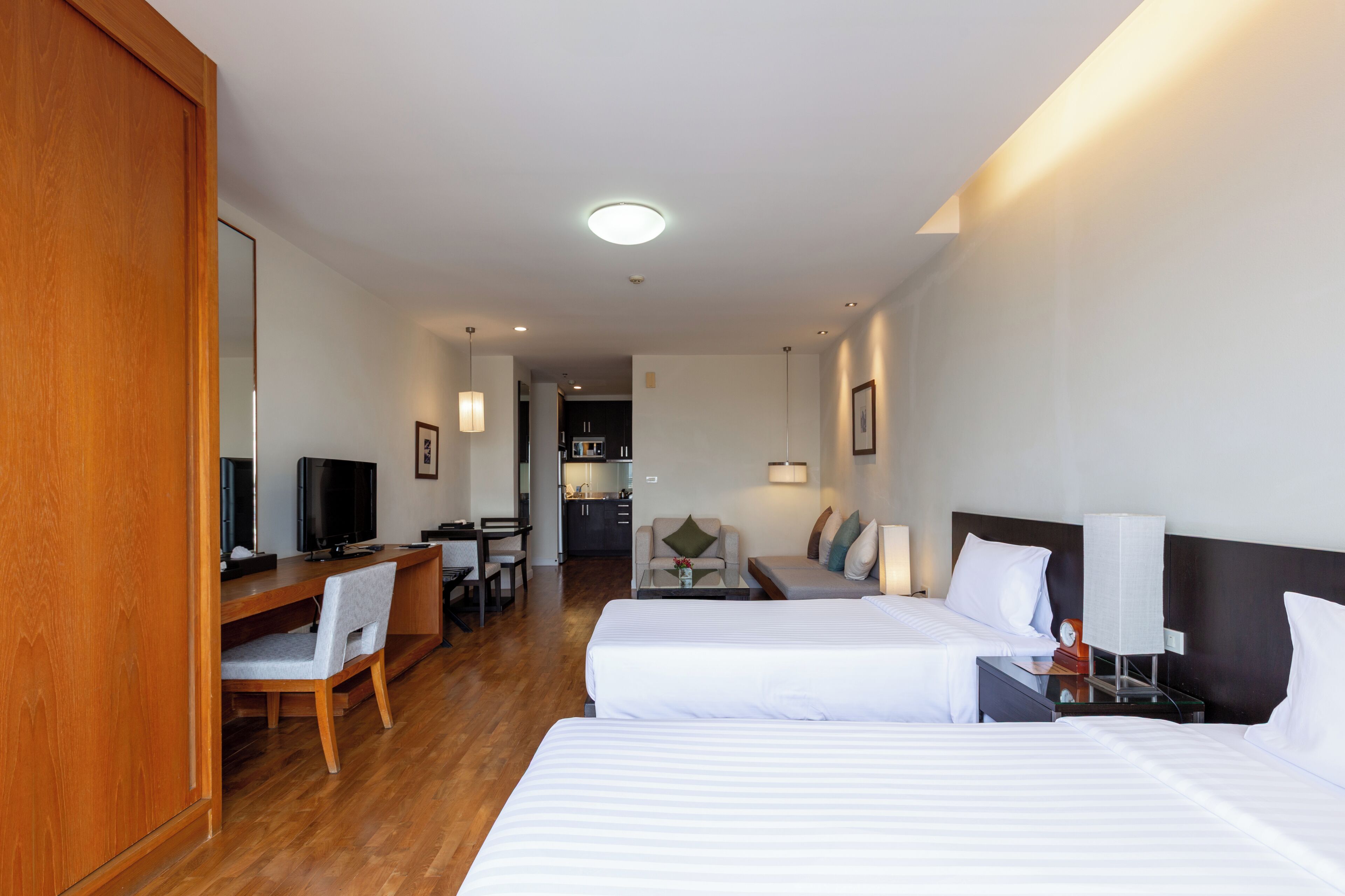 A hotel room with two beds, a desk with a chair, a television, and a kitchenette in the background.