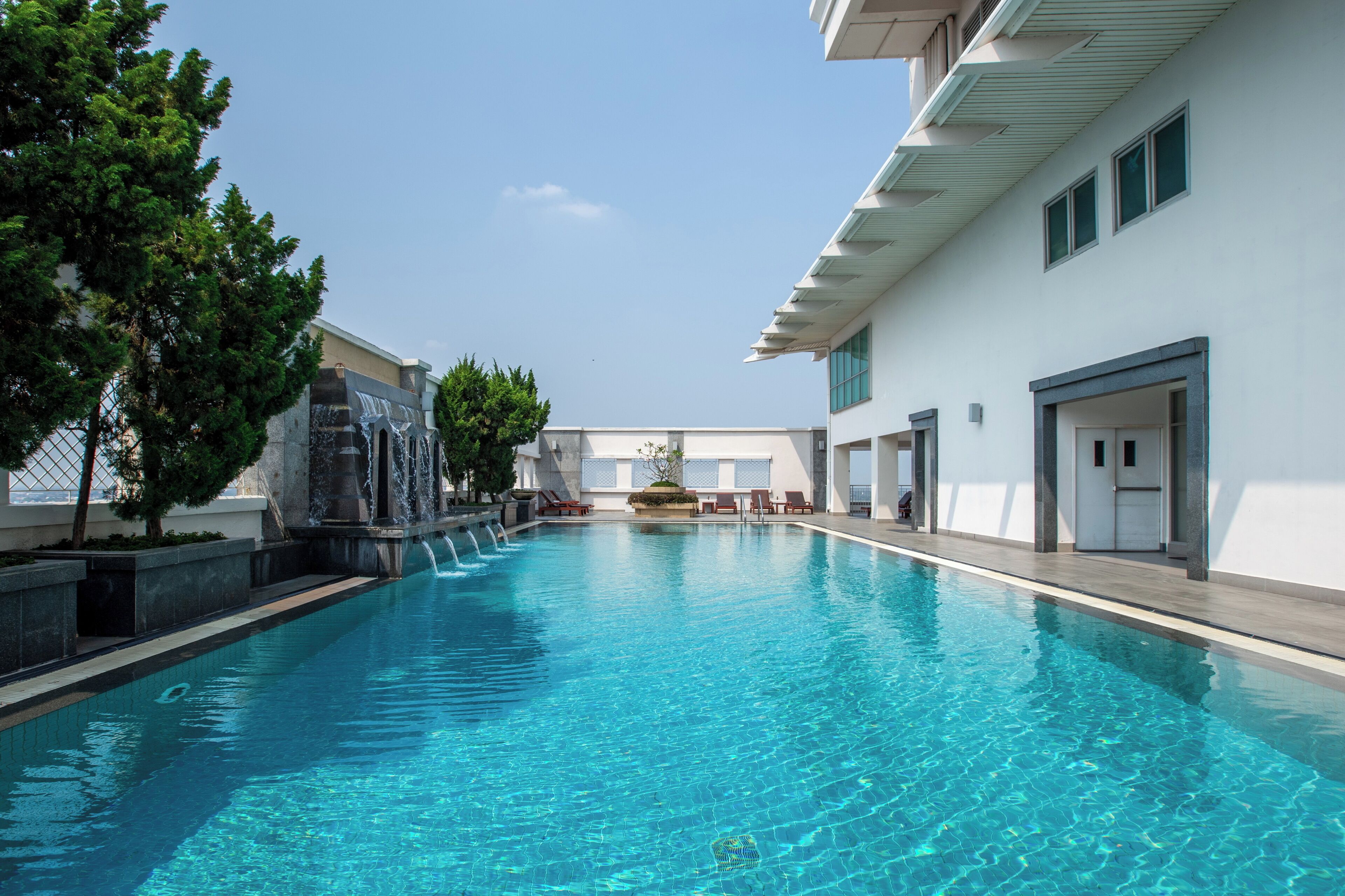 A swimming pool with a waterfall feature, surrounded by trees and a building with a modern design.