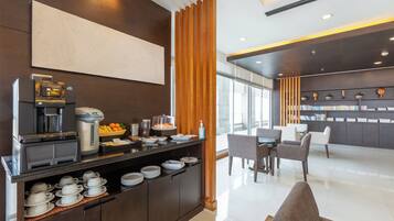 A modern hotel room with a breakfast bar, coffee machine, fruit bowl, and a seating area with a table and chairs.