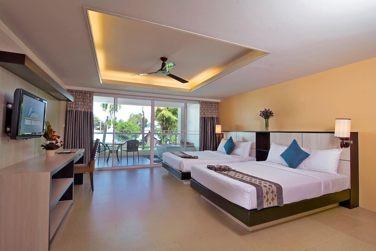 A hotel room with two beds, a desk, a TV, and a balcony with a view of greenery.