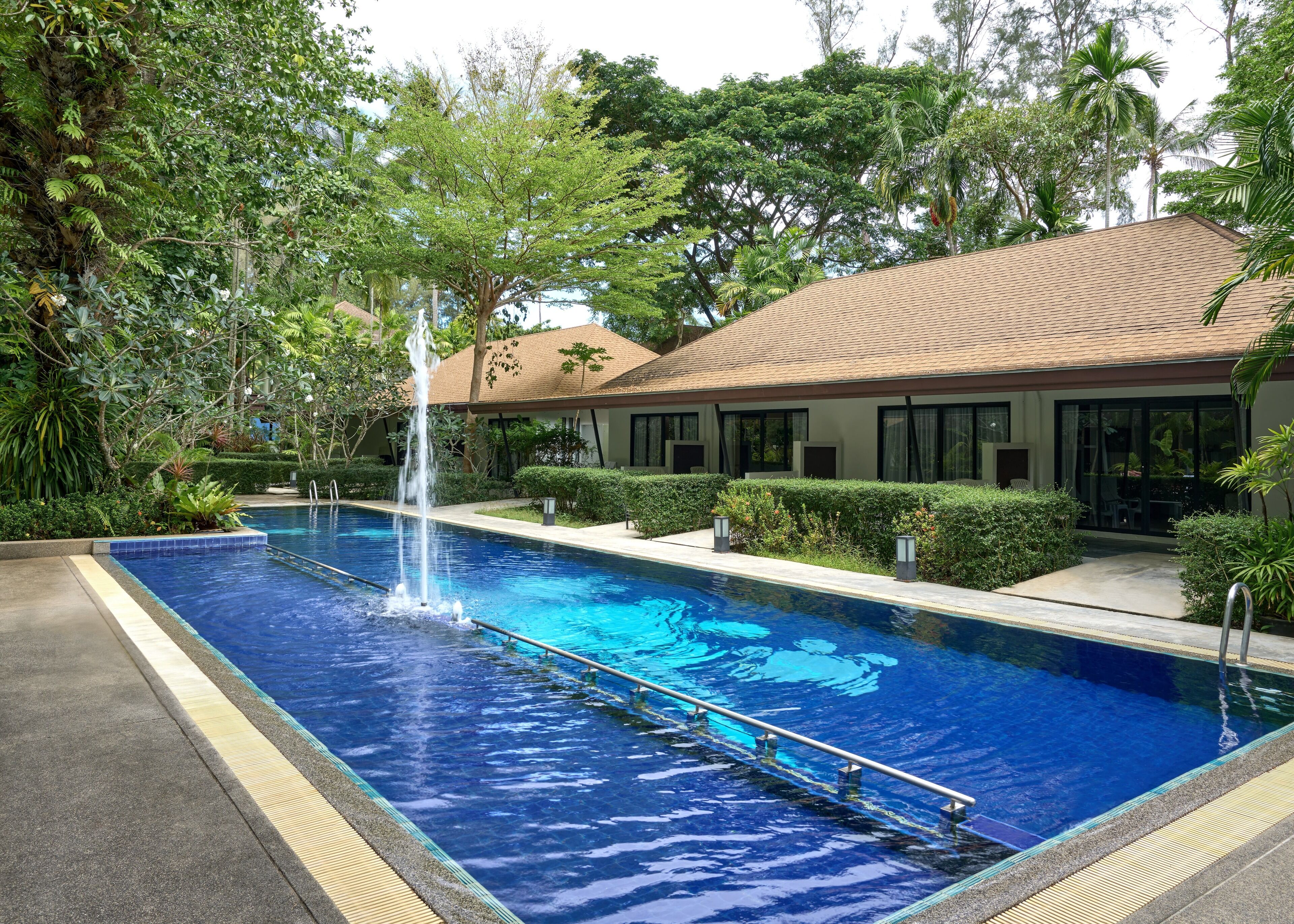 Une piscine avec une fontaine, entourée d’une végétation luxuriante et bordée d’un bâtiment dont le toit est recouvert de tuiles.
