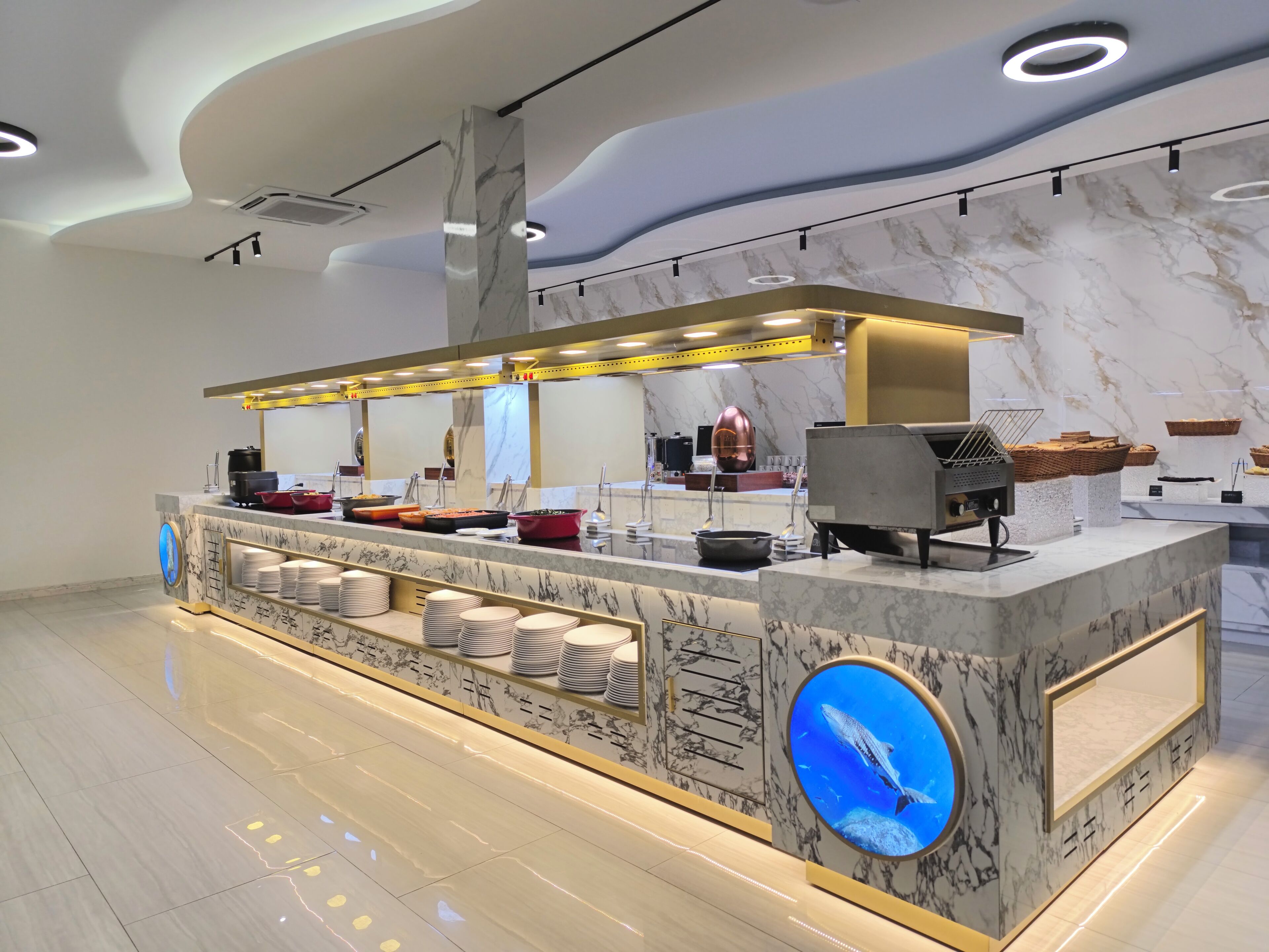 Een moderne buffetopstelling met marmeren aanrechtbladen, een centraal eiland met etalages en een groot rond aquarium.