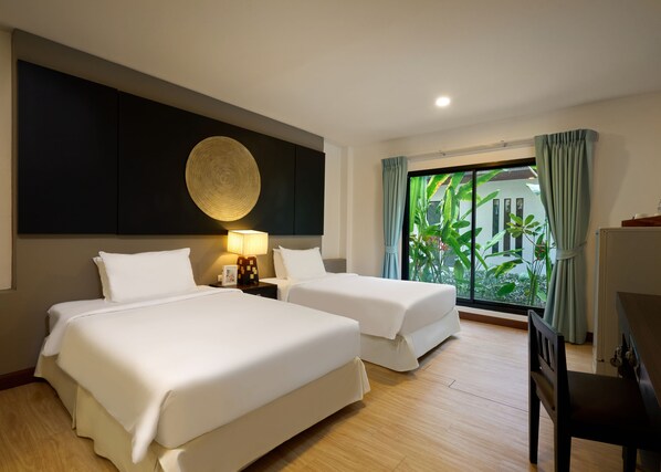 Habitación de hotel con dos camas, un adorno circular grande en la pared, una mesita de noche con lámpara y vistas a zonas verdes a través de una ventana.