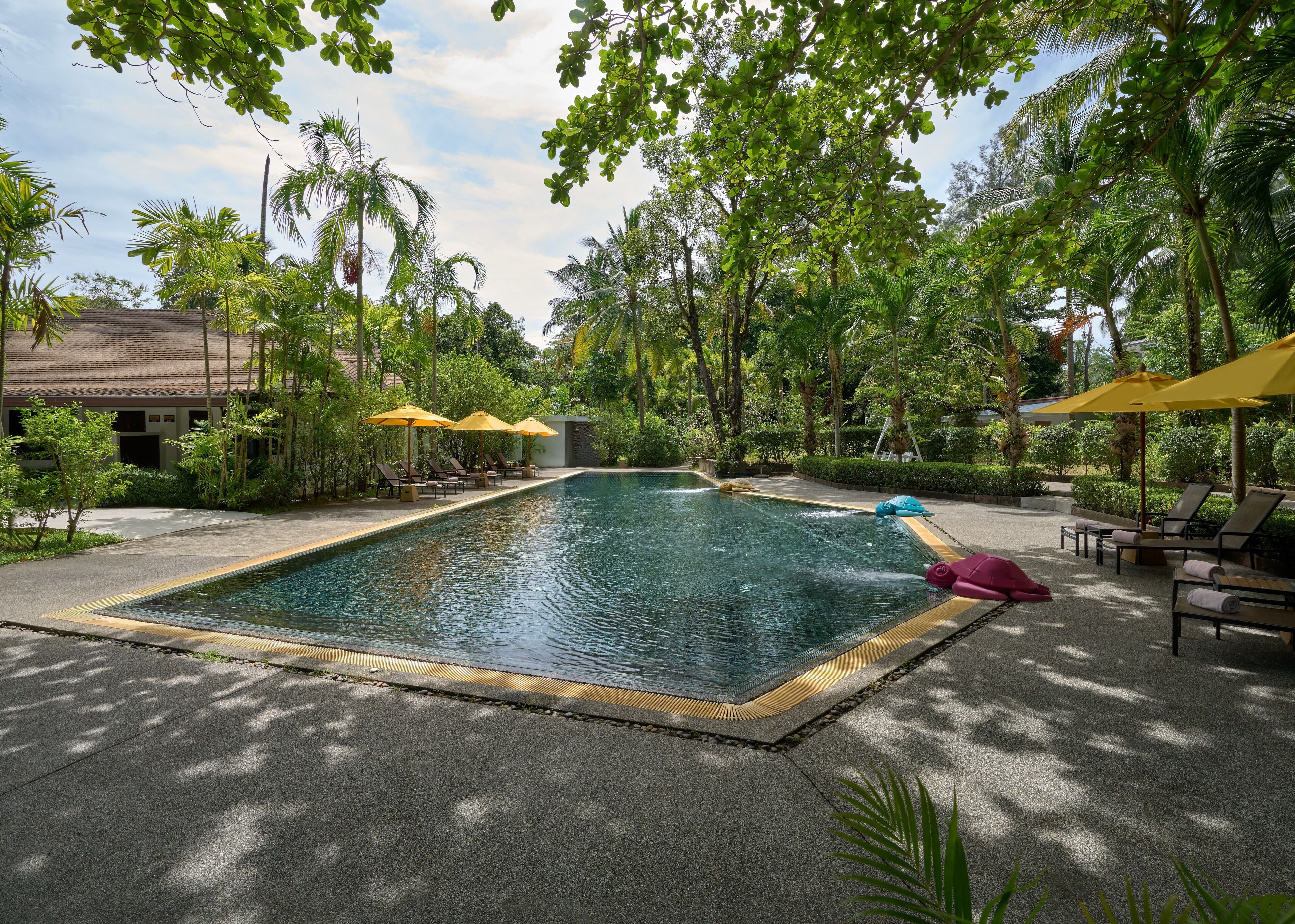 Une piscine entourée de chaises longues et de parasols, le tout baigné d’une végétation luxuriante et bordé de palmiers.