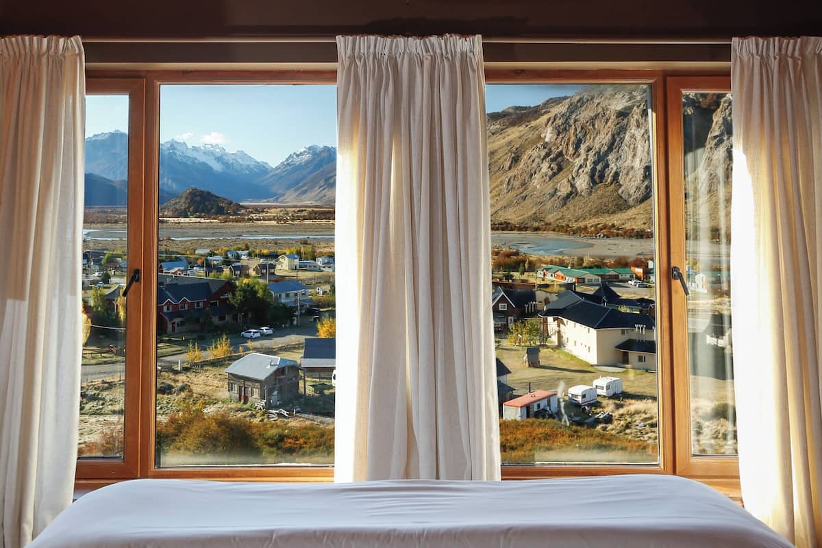 A bedroom with a large bed, wooden ceiling, and a view of a town and mountains through a large window.