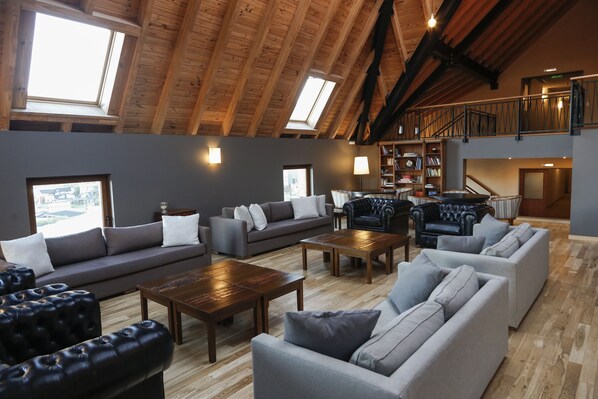 A spacious loft-style living area with high ceilings, wooden beams, and skylights. It features a gray sofa set, a black leather armchair, and a wooden coffee table.