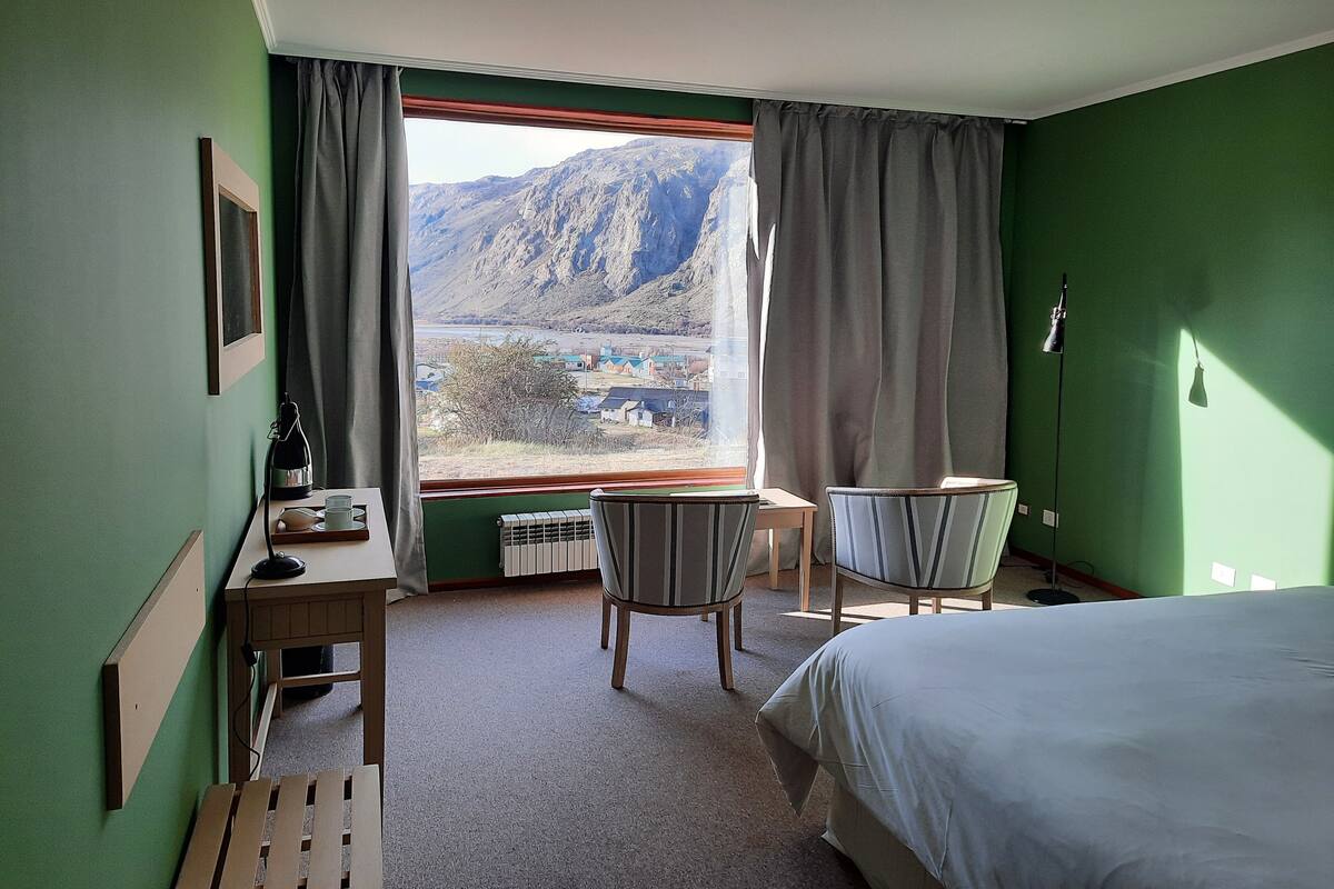 A hotel room with a bed, two chairs, a desk with a lamp, a radiator, and a view of mountains through the window.