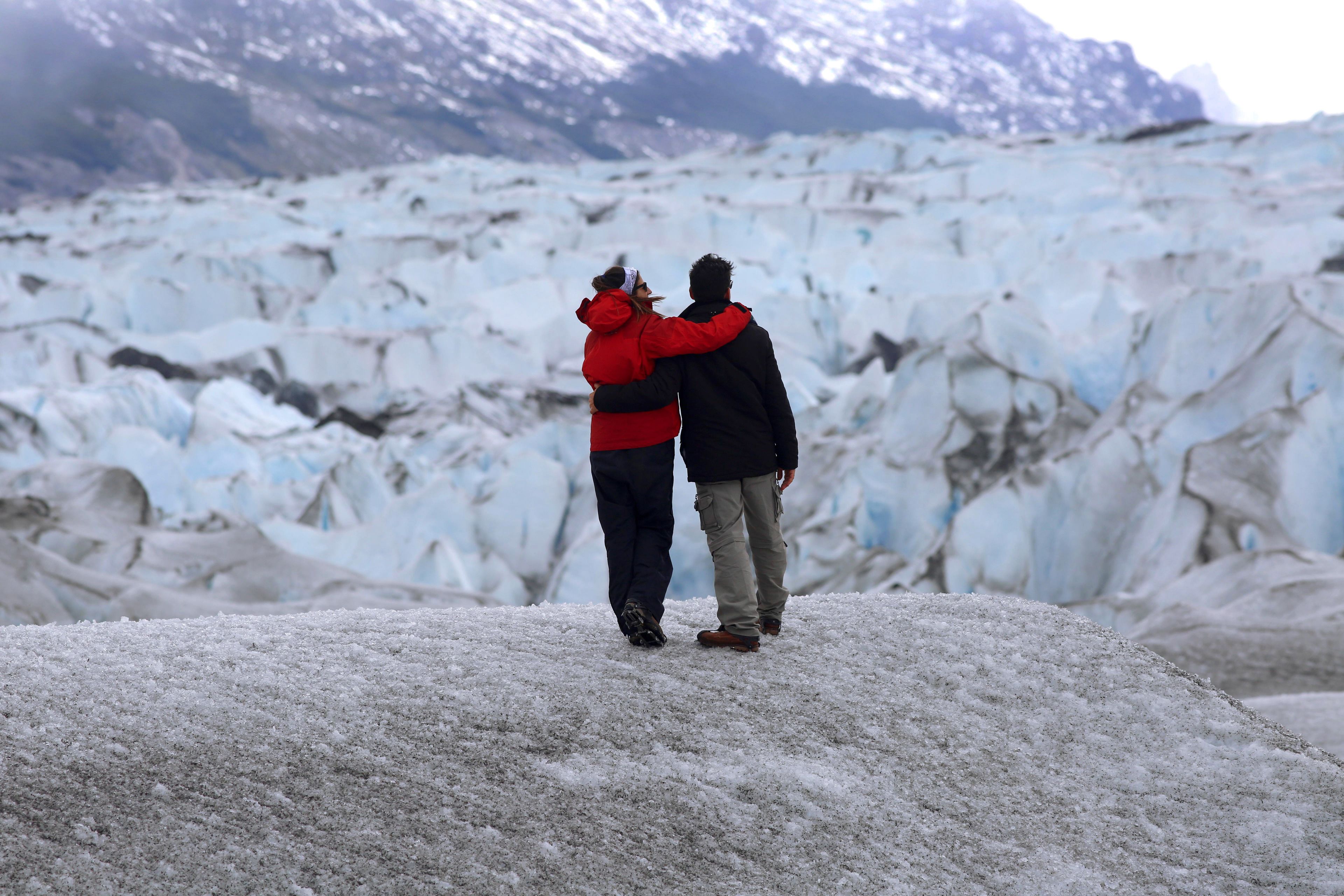 Duas pessoas em pé numa encosta coberta de neve, com uma geleira ao fundo.