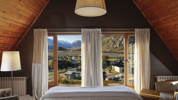 A bedroom with a large bed, a chair, a table, and a view of a village and mountains through large windows. - Los Cerros del Chalten Boutique Hotel (El Chalten)
