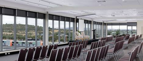 Un salón de conferencias con filas de sillas, ventanas amplias y un escenario con un atril.
